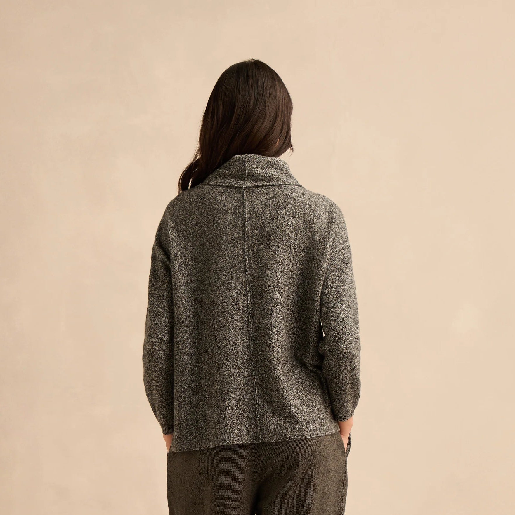 A woman is shown from behind wearing a textured gray knit cowl-neck sweater and dark olive green wide-leg cropped trousers, paired with brown slip-on shoes, against a solid beige studio background.