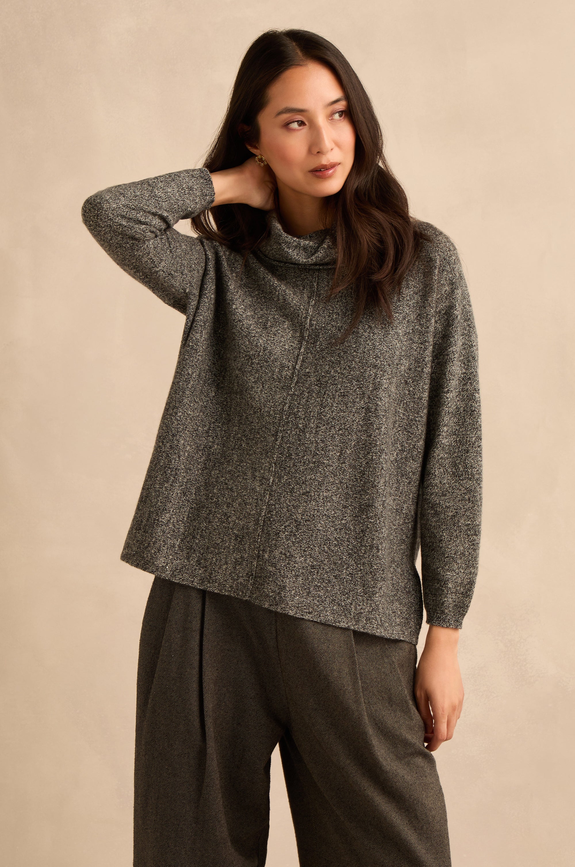 A woman wearing a textured charcoal gray mock-neck knit sweater and matching dark gray trousers, posing against a neutral beige studio backdrop.