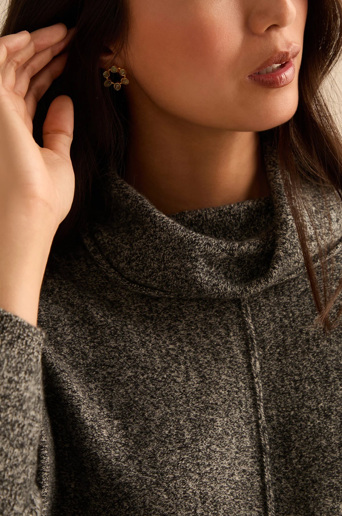 Close-up of a woman wearing a dark gray heathered cowl-neck sweater and a small gold flower-shaped earring, with her hand near her hair.