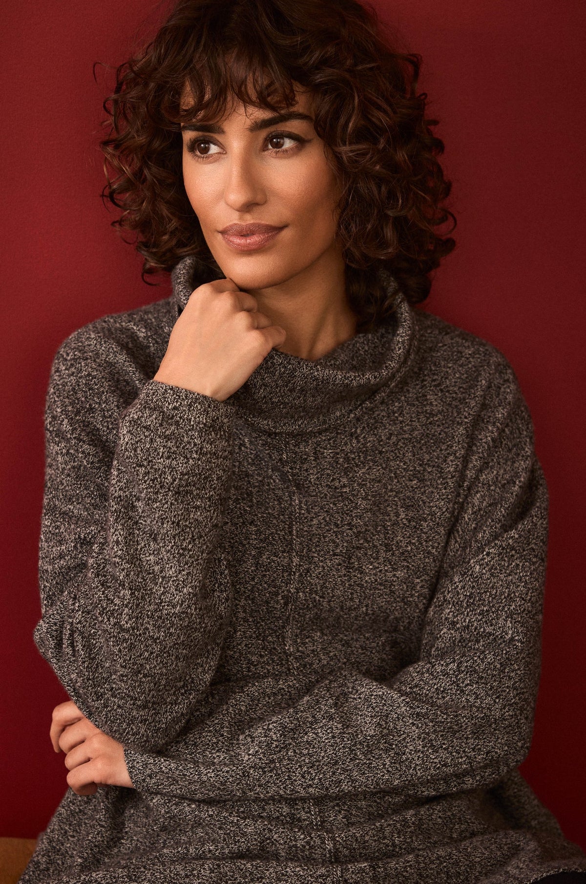 A woman with curly brown hair wearing a dark gray marled turtleneck sweater, posing against a solid dark red background.