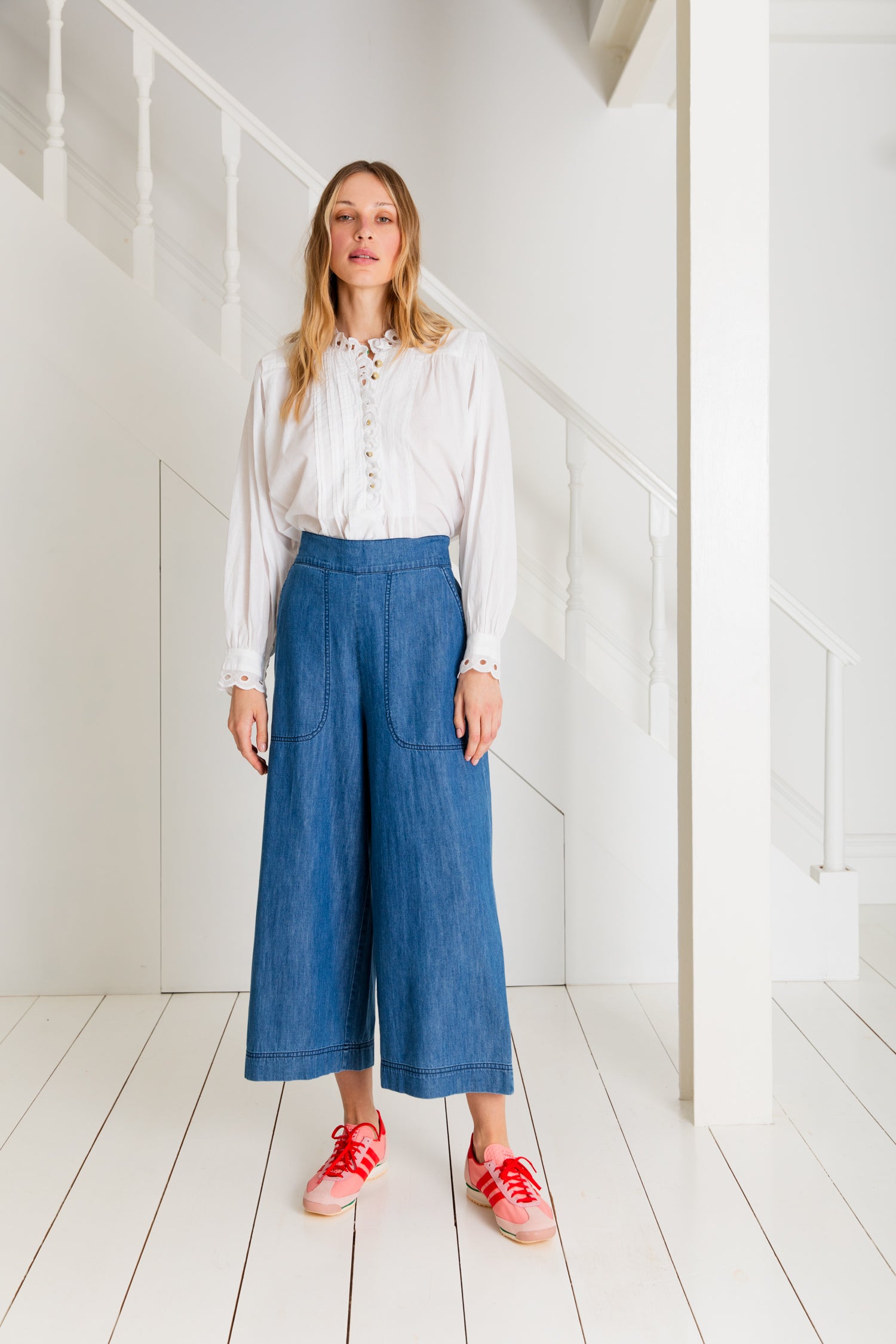 Woman wearing a white eyelet blouse and wide-leg denim culottes.  She is wearing pink sneakers with red laces.