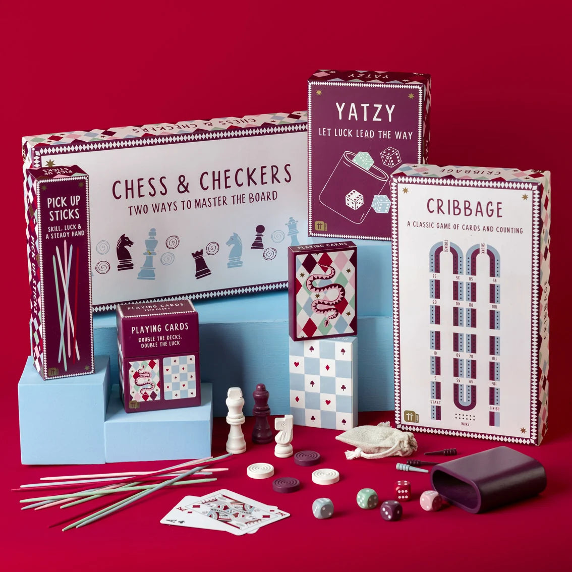 A boxed card game set displayed on a red background, including Chess & Checkers, Cribbage, and a purple box labeled Yatzy, with game components such as cards, pawns, dice, checkers, and wooden sticks arranged in front.