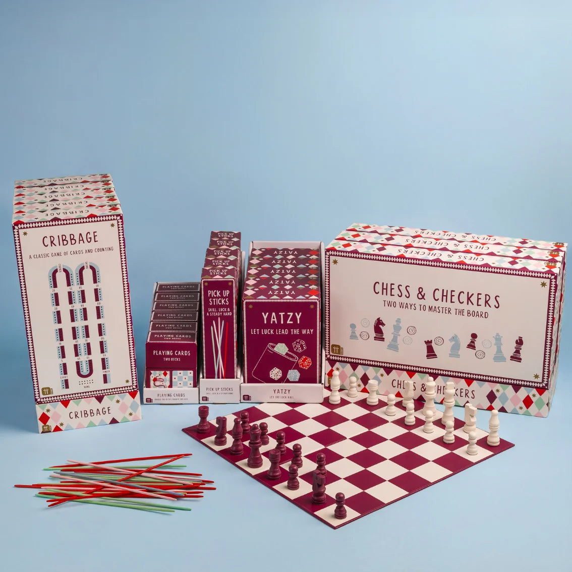 A children's chess game set displayed on a light blue background, including multiple branded boxes, stacks of pieces, a chess board, and colored sticks. Visible products include Cribbage, Chess & Checkers, and Yatzy.