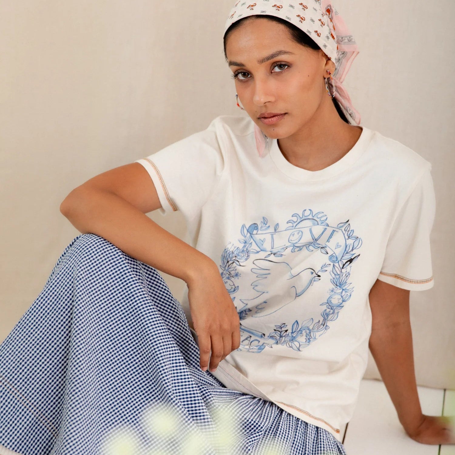Woman wearing a white t-shirt with a graphic design and a headscarf, sitting on a white surface.