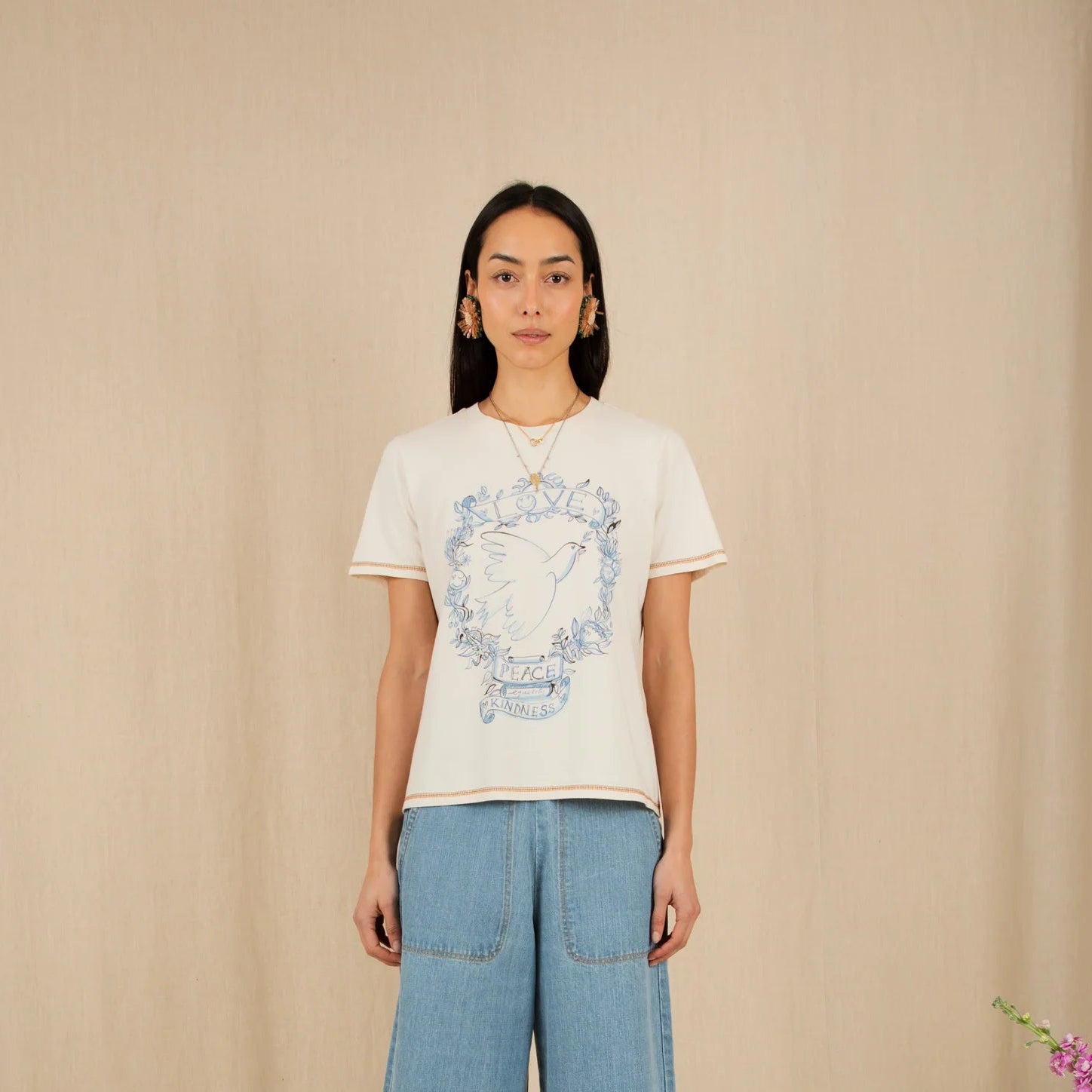 Person wearing a white t-shirt with a graphic design and blue jeans standing against a beige wall with flowers on either side.