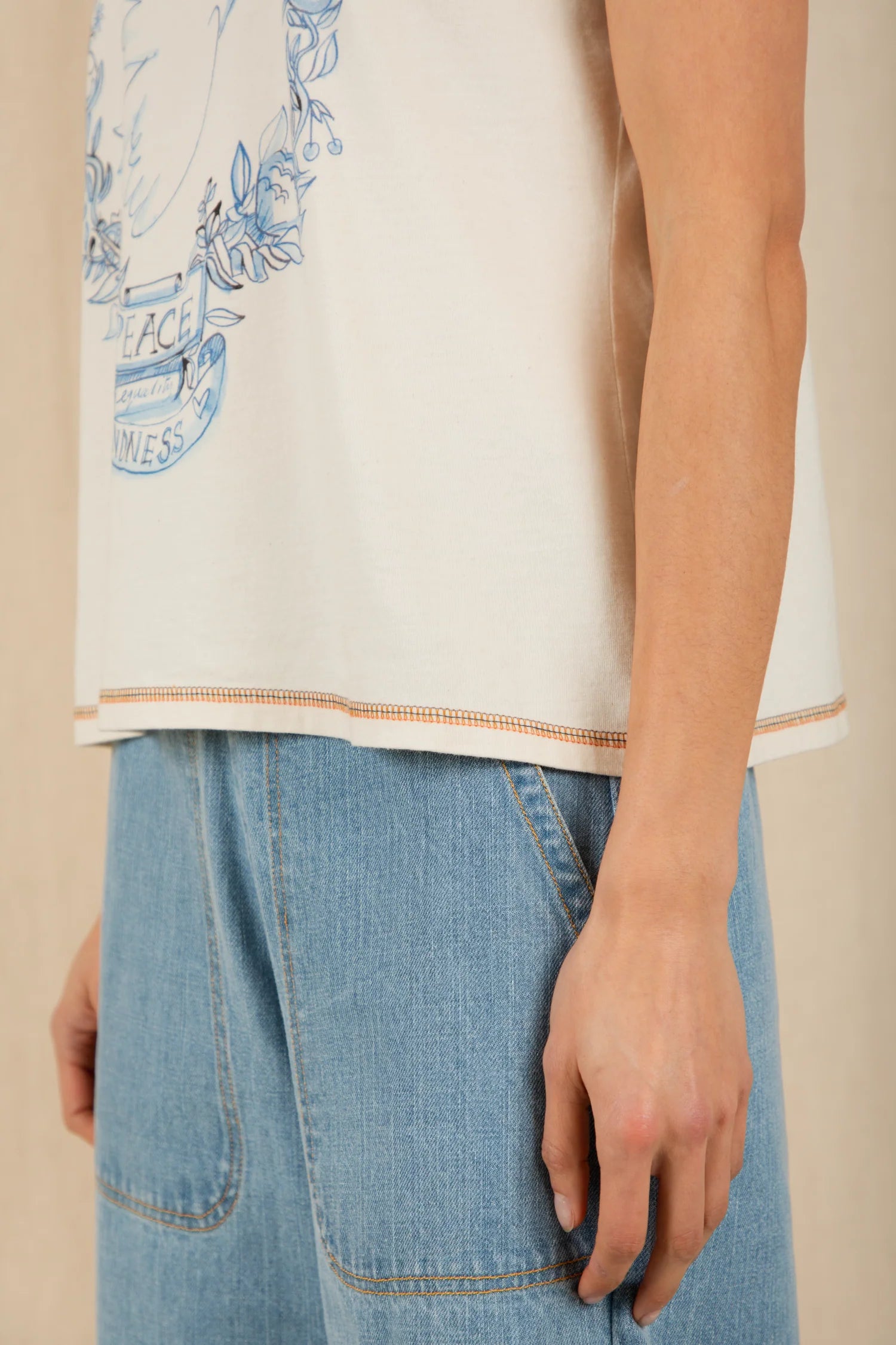 Person wearing a white t-shirt with blue graphics and text, paired with light blue jeans on a beige background.