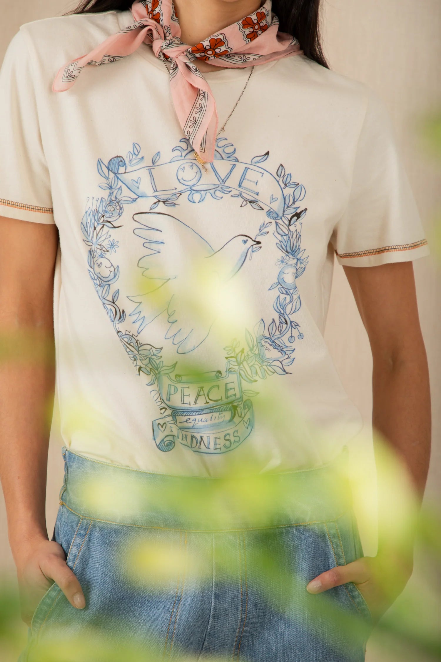 Person wearing a white t-shirt with a graphic design and a pink scarf, standing in front of green foliage.