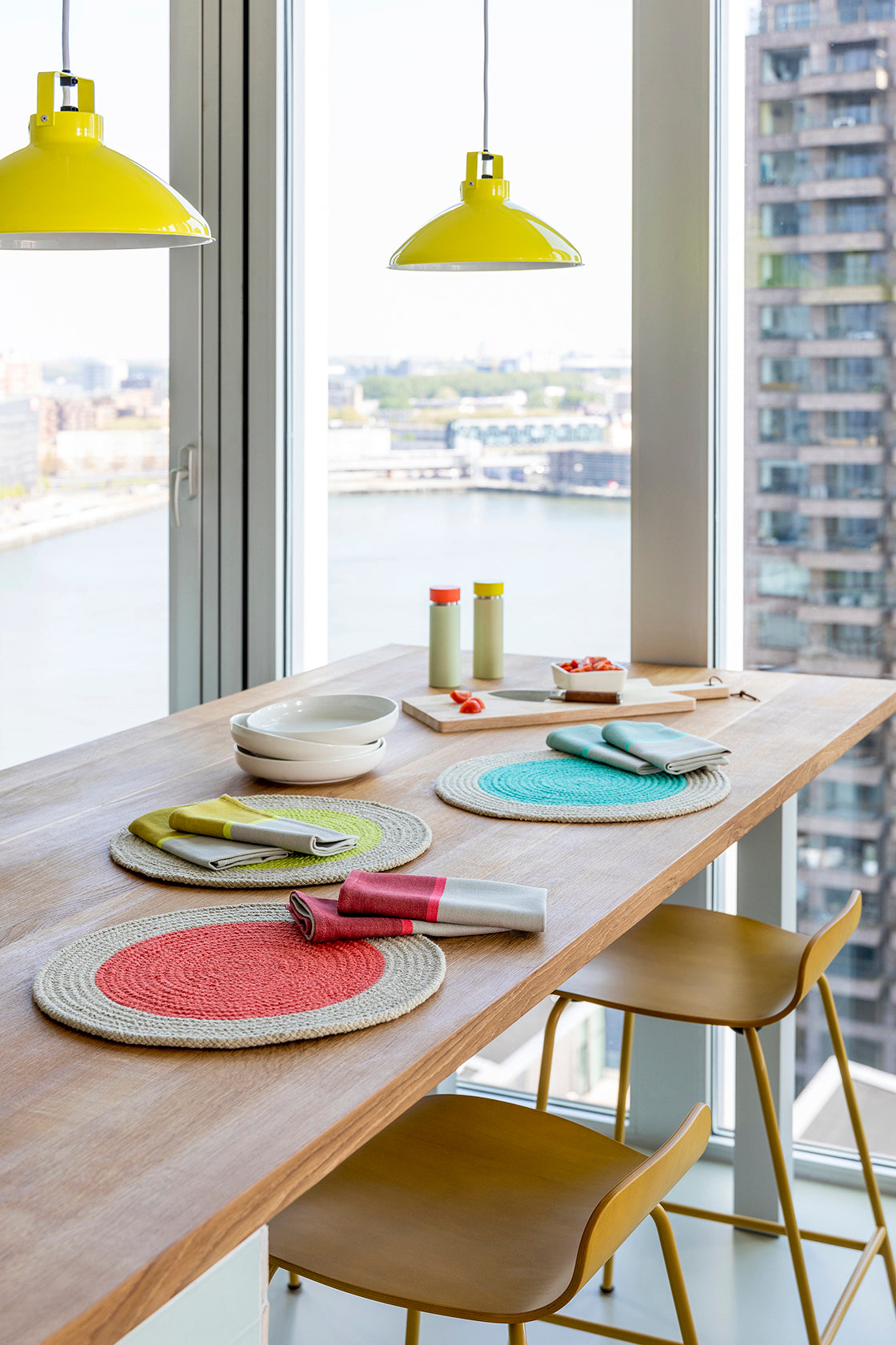 Three round jute placemats in coral, teal, and lime green, with folded napkins on a light wood table.