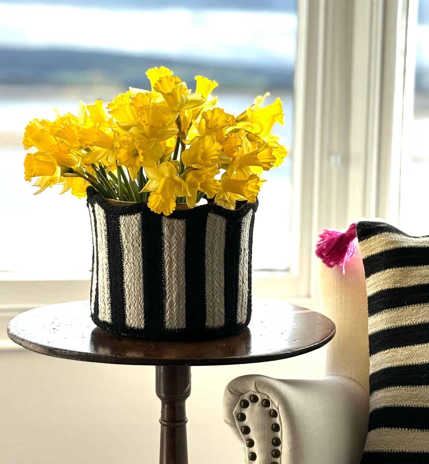 Black and white striped woven basket filled with a bouquet of yellow daffodils, sitting on a dark wood side table.