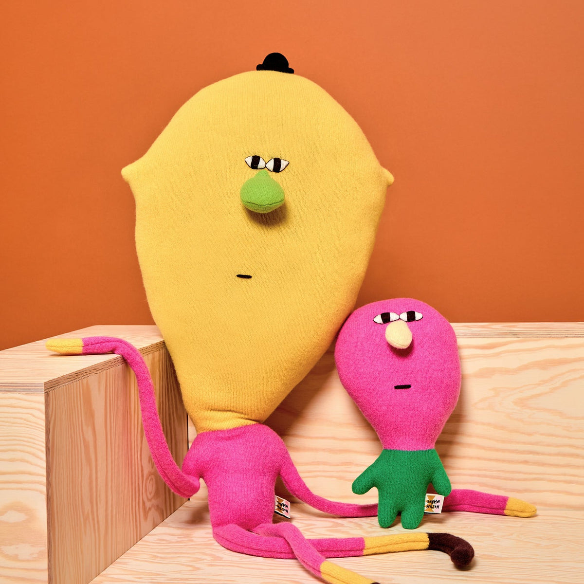 Two colorful plush toys resembling a yellow lemon and pink creature on a wooden surface with an orange background.