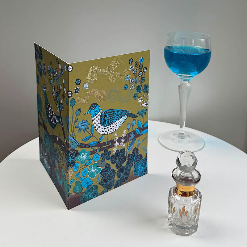 Decorative card with a bird design, glass with blue liquid, and perfume bottle on a white surface.