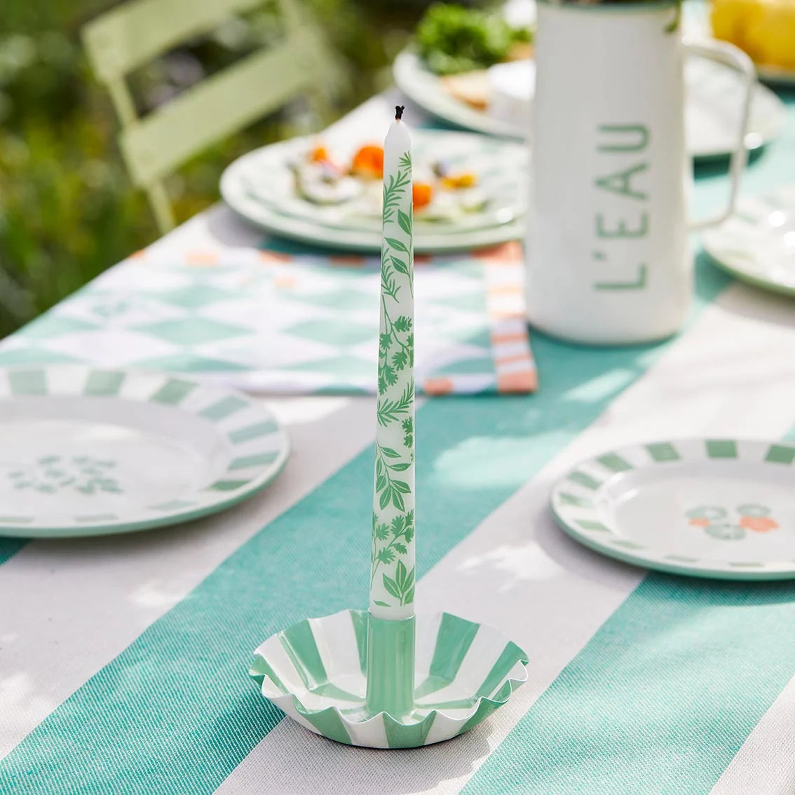 Green and white decorative candle on a striped tablecloth with plates and a mug in the background.