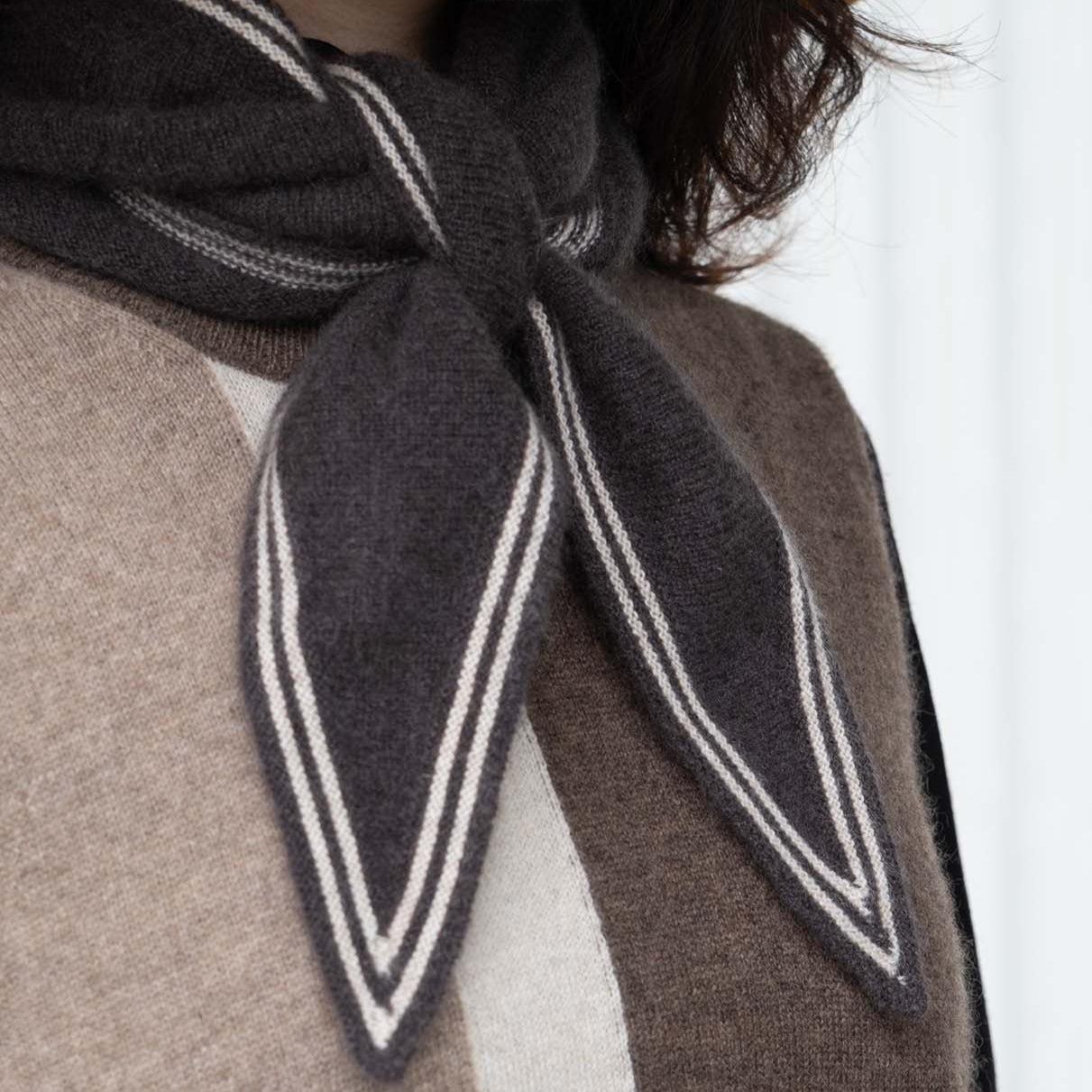 Person wearing a dark gray scarf with white stripes over a brown garment.