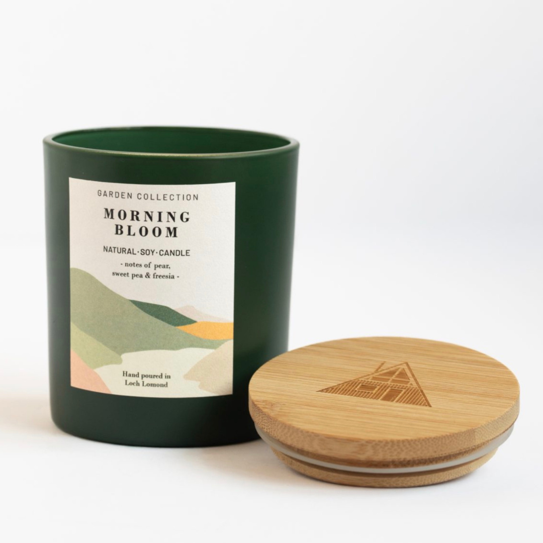 A dark green soy candle jar with a white label reading 'Garden Collection Morning Bloom' and a light natural bamboo lid placed beside it on a clean white background.