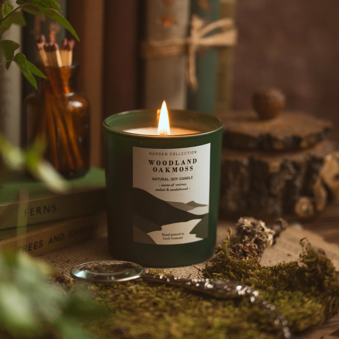 A lit dark green candle in a cylindrical jar sits on a rustic surface with burlap, moss, and wood props. The front label reads 'Garden Collection Woodland Oakmoss Natural Soy Candle'.