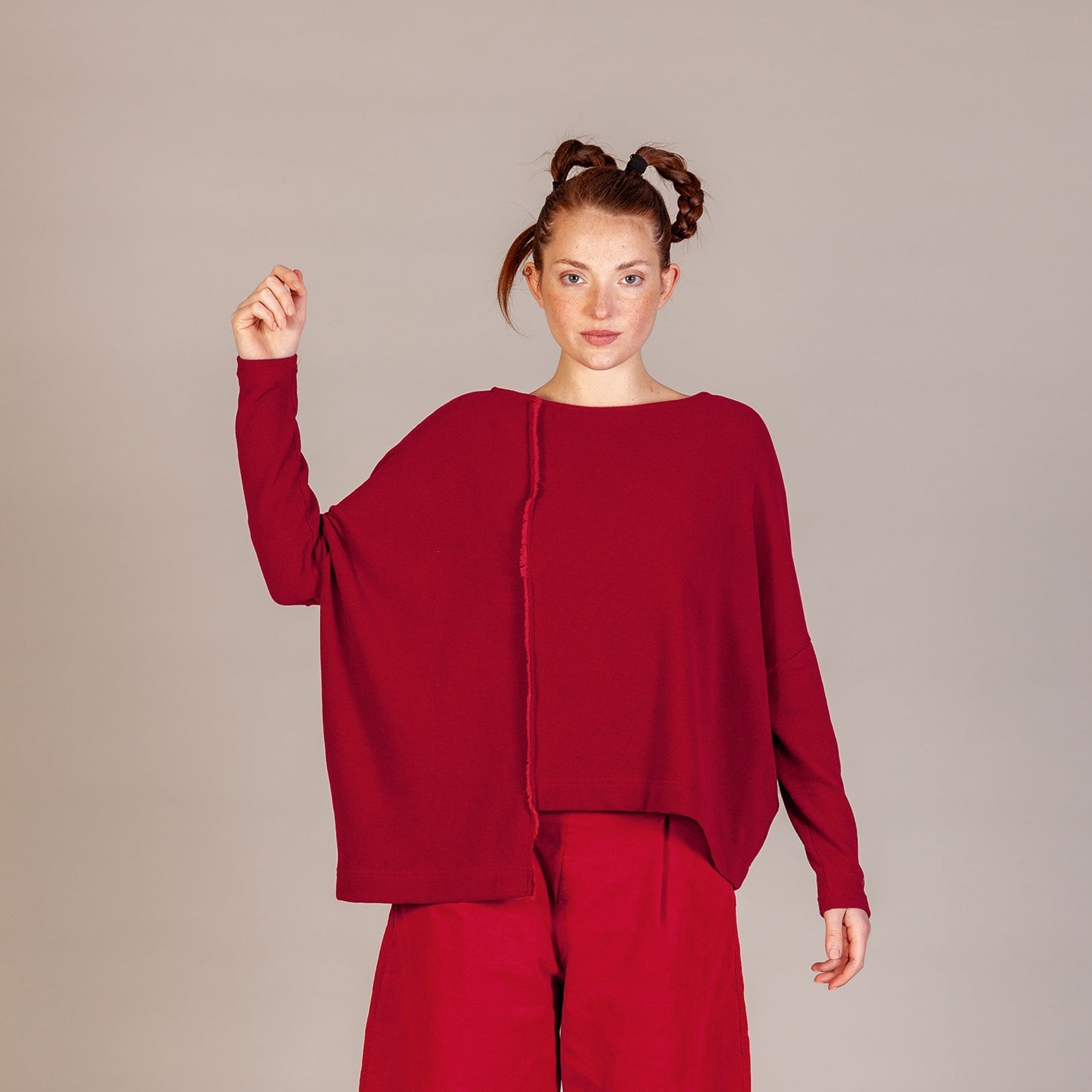 A model stands against a neutral light gray studio background wearing a matching deep red two-piece outfit consisting of a loose, oversized long-sleeve top and wide-leg cropped pants, paired with black shoes.