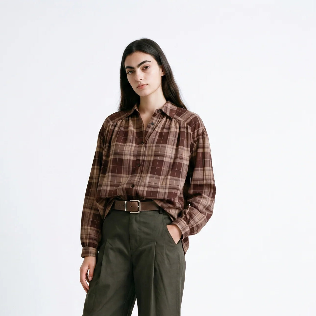 A woman stands against a light gray studio background wearing a brown and burgundy plaid button-up shirt, olive green wide-leg trousers, a brown belt, and dark brown loafers.