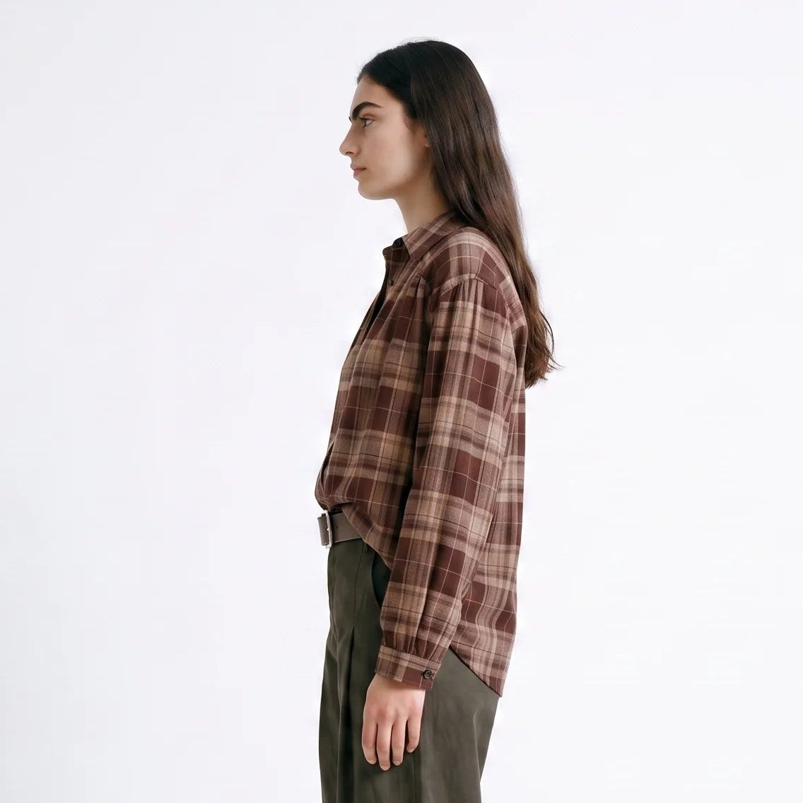 A woman stands in profile wearing a brown and beige plaid button-up shirt, olive green wide-leg trousers, and dark brown loafers against a plain white studio background.