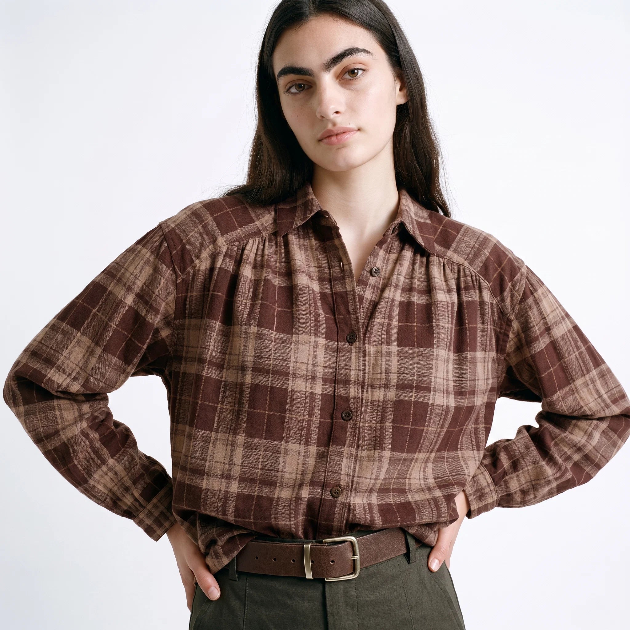 A woman modeling a brown and beige plaid long-sleeve button-up shirt with a gathered, ruffled neckline, paired with dark green trousers and a brown belt, against a plain white studio background.
