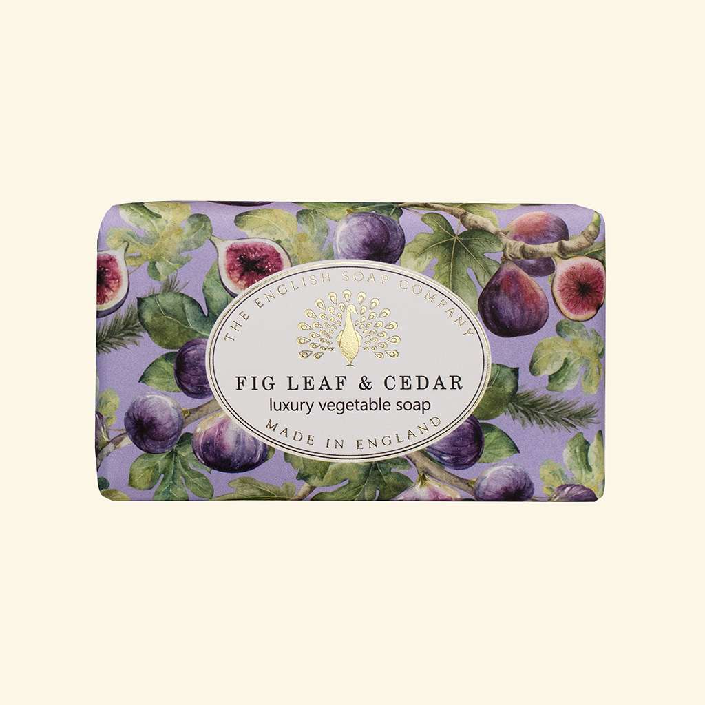 A rectangular bar of soap wrapped in a lavender illustration pattern featuring fig leaves, purple figs, and green foliage, with a central white oval label reading 'The English Soap Company Fig Leaf & Cedar luxury vegetable soap Made in England'.