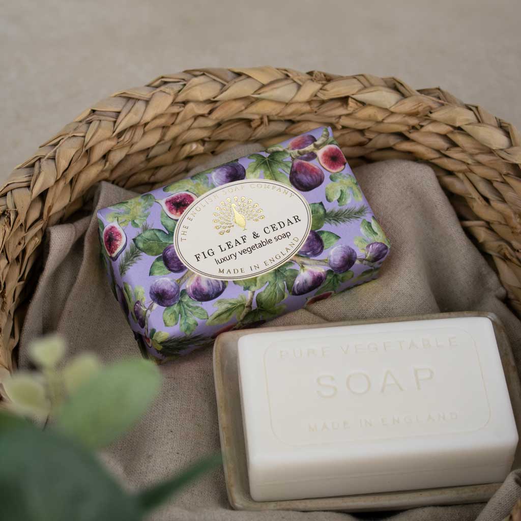 Two bars of soap displayed in a woven wicker basket lined with beige fabric. The soap in the front is an off-white bar labeled 'PURE VEGETABLE SOAP', and the soap behind it is wrapped in a lavender paper sleeve printed with purple figs and green leaves, labeled 'FIG LEAF & CEDAR'.