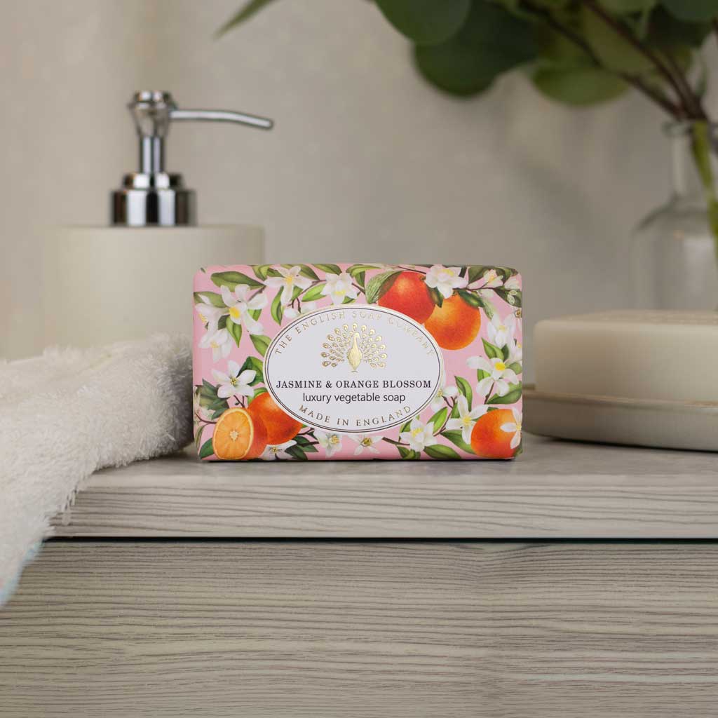 A rectangular bar of luxury vegetable soap in a pink floral wrapper labeled 'Jasmine & Orange Blossom' sits on a gray wooden surface. The wrapper features illustrations of white blossoms and bright orange citrus fruits.