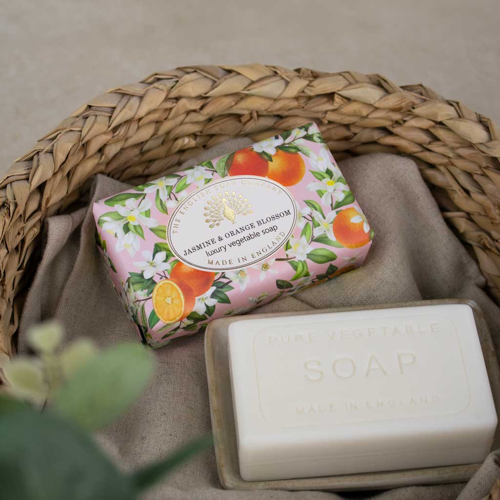 A packaged bar of soap and an unwrapped bar of soap displayed in a woven basket on a neutral fabric surface. The wrapped soap has a pink floral design with orange and white blossoms, and the label reads Jasmine & Orange Blossom Luxury Vegetable Soap, Made in England.