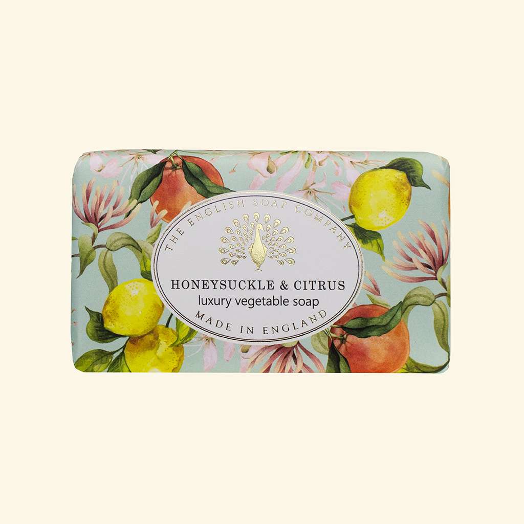 A rectangular bar of soap wrapped in decorative paper featuring lemons, peaches, and pink flowers on a pale blue-green background, with a white oval label reading 'The English Soap Company' and 'Honeysuckle & Citrus luxury vegetable soap Made in England'.