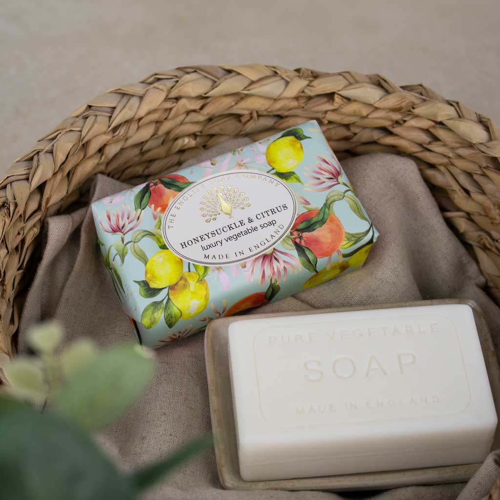 A packaged bar of soap and an unwrapped bar of soap displayed in a woven basket on a neutral fabric surface. The wrapped soap has a pastel floral and citrus print reading 'HONEYJUNKLE & CITRUS luxury vegetable soap MADE IN ENGLAND,' and the unwrapped soap is plain white with embossed text reading 'PURE VEGETABLE SOAP MADE IN ENGLAND.