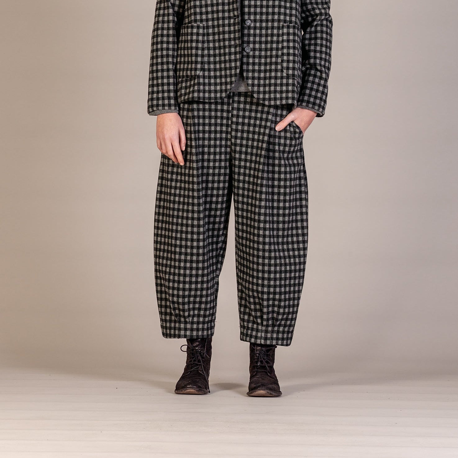 A model stands against a neutral studio backdrop wearing a matching black and white checkered blazer and trousers set, styled with a gray turtleneck and black boots.