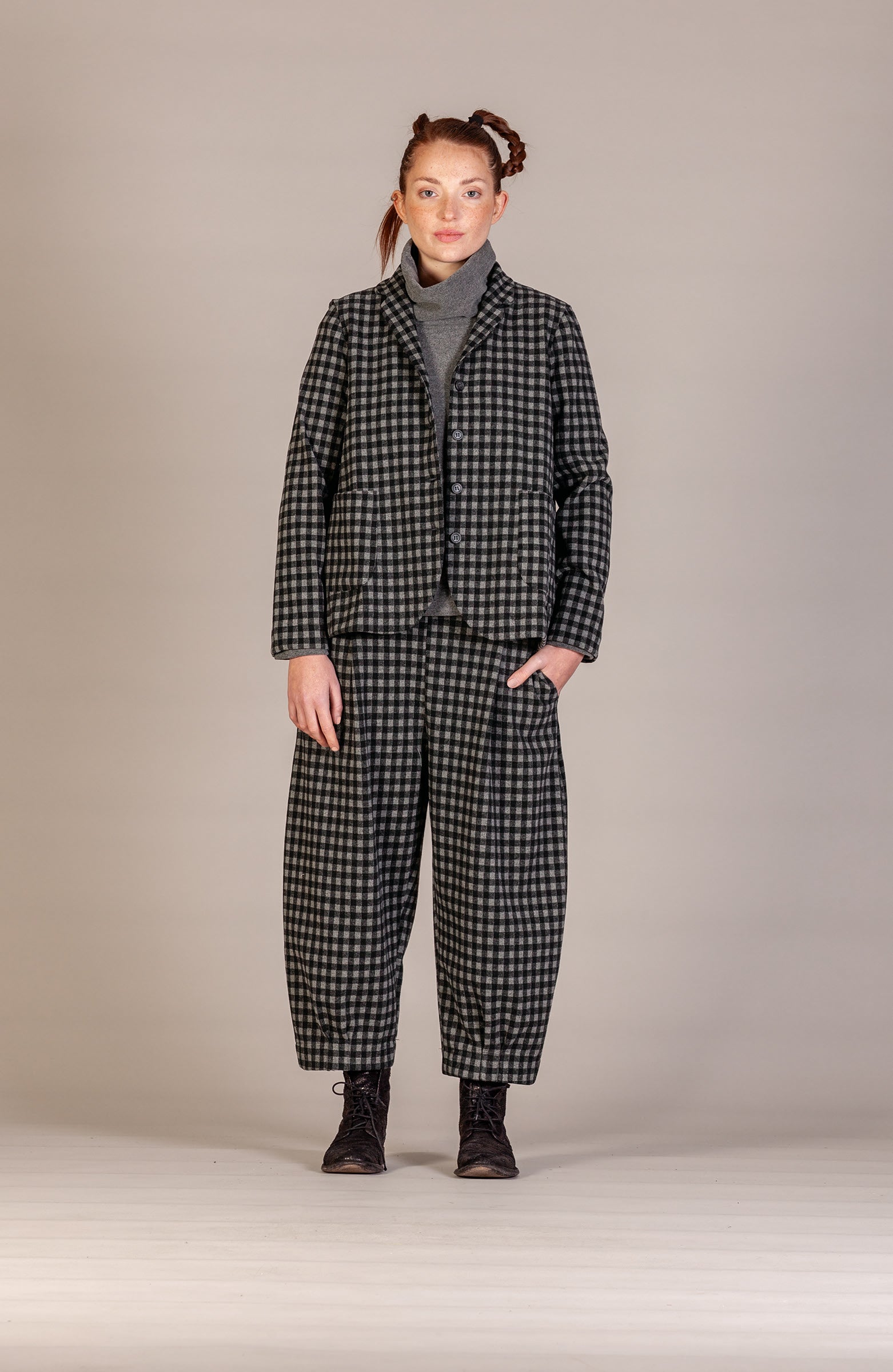 A model stands against a neutral studio backdrop wearing a matching black and white checkered blazer and trousers set, styled with a gray turtleneck and black boots.