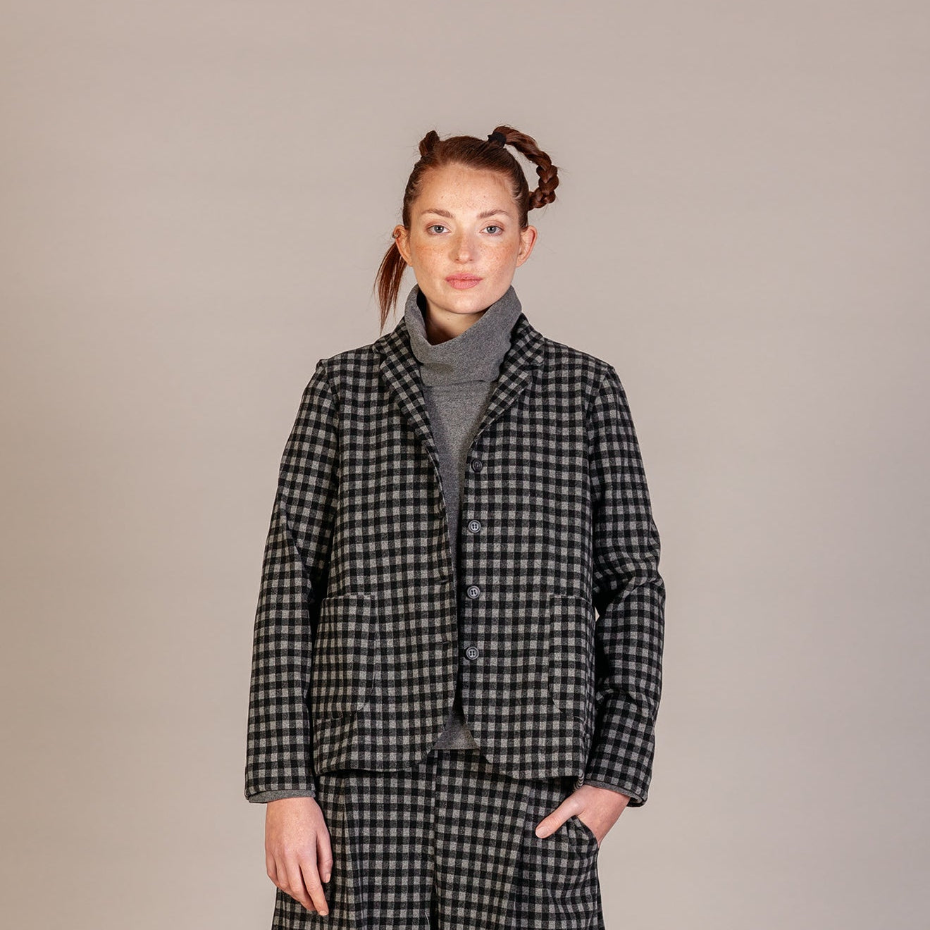 A model stands against a neutral studio backdrop wearing a matching black and white checkered blazer and trousers set, styled with a gray turtleneck and black boots.