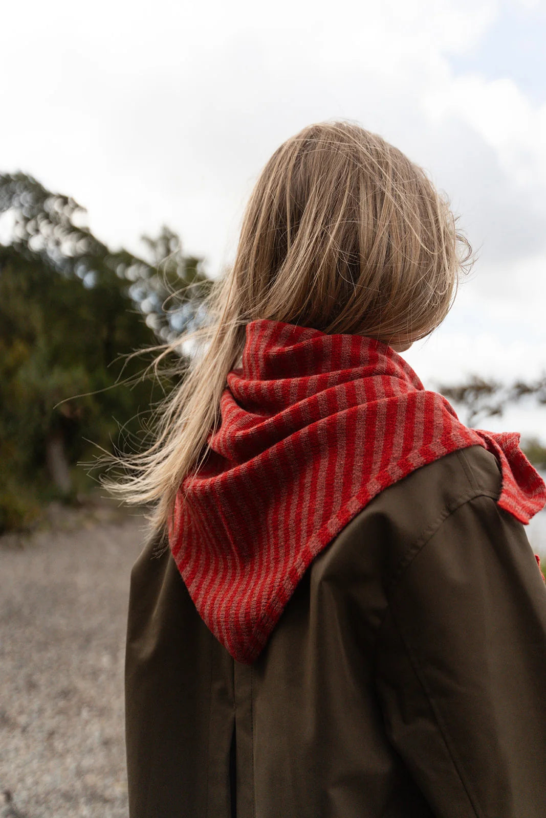 A person viewed from behind wearing a red and brown striped knit scarf and a brown coat outdoors under a cloudy sky.