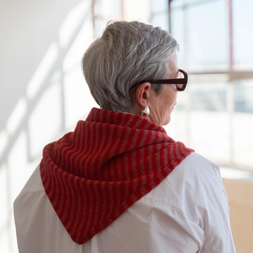 A person with short gray hair and black glasses, seen from behind, wearing a red and white striped knit hooded scarf or shawl over a white shirt in a bright indoor setting.