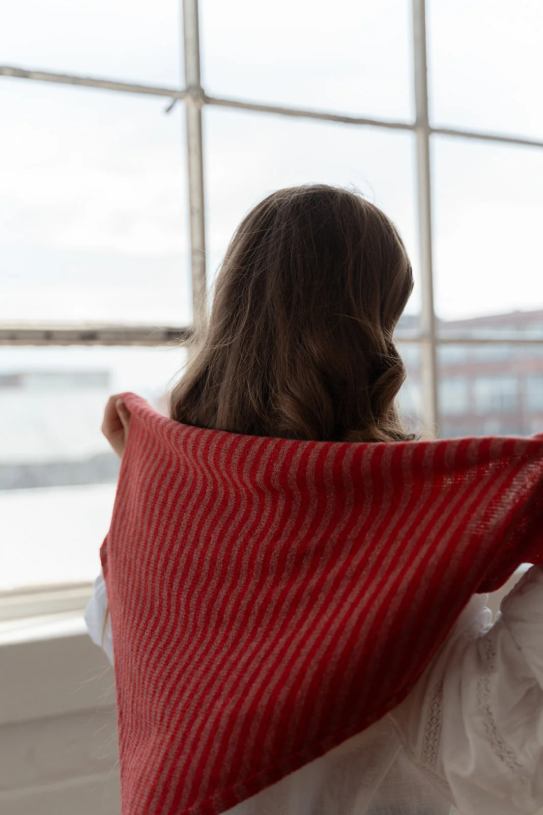 A woman viewed from behind wearing a red and white striped knit shawl draped over her shoulders, standing in front of a large industrial-style window.