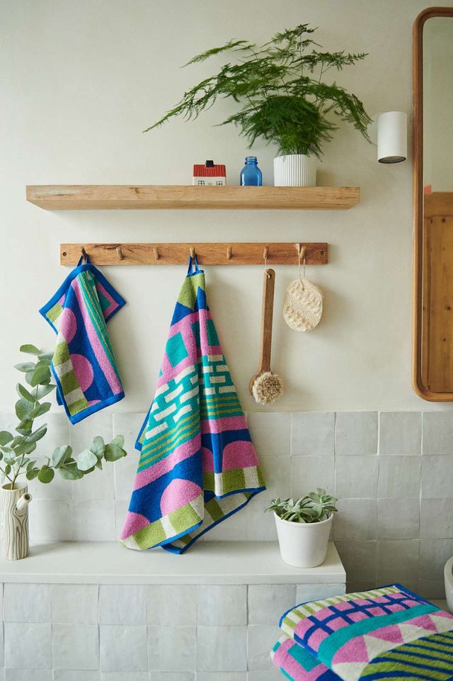 Colorful geometric patterned bath towels hanging on a wooden wall hook rack in a bright bathroom setting, with a wooden floating shelf, potted plants, and a tiled wall in the background.