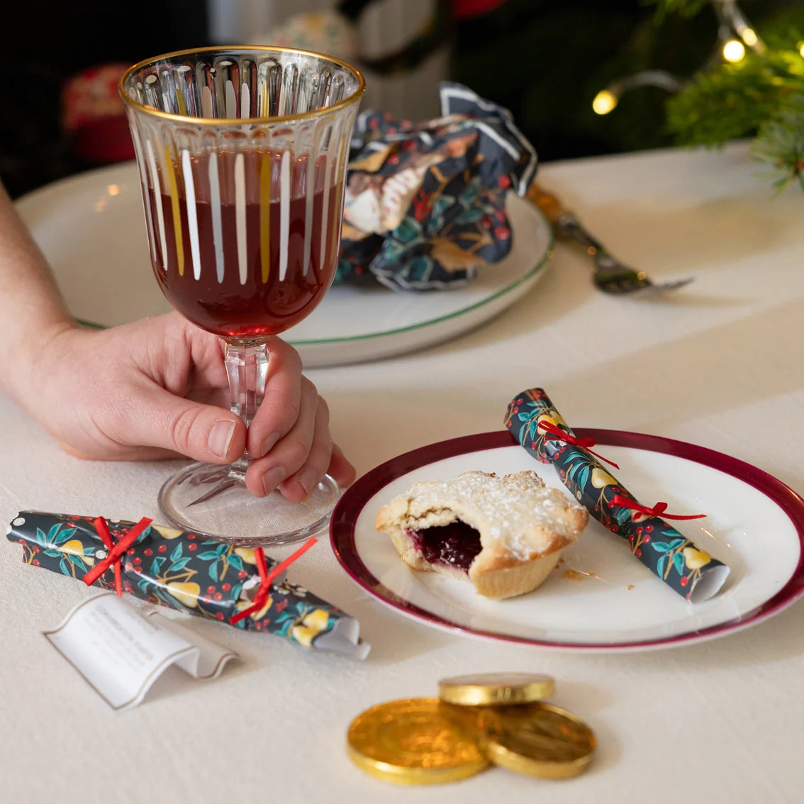 Hand holding a glass of red wine with a plate of mince pie and gold coins on a festive table.