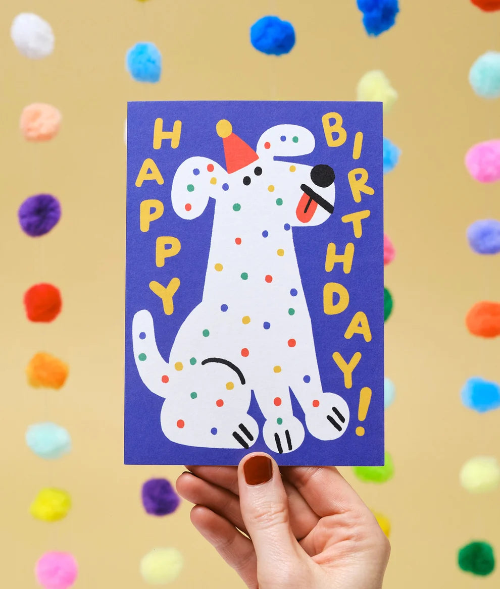 Hand holding a birthday card with a polka dot dog illustration against a colorful pom-pom background.
