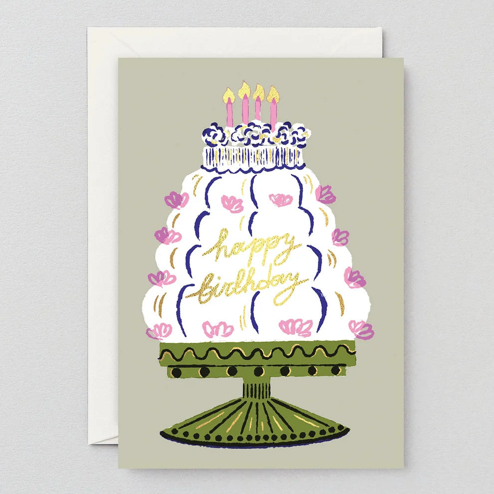 A square birthday greeting card with a white envelope behind it, featuring an illustrated white cake on a stand with pink flowers, blue piping, yellow candles, and gold 'happy birthday' script on a light warm gray background.
