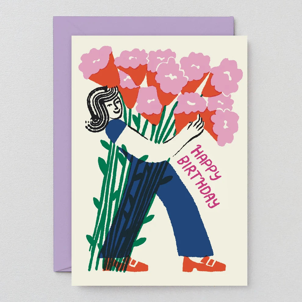 A square birthday greeting card with a lavender envelope behind it, featuring a stylized illustration of a woman holding a large bouquet of pink flowers and green leaves, with the words “HAPPY BIRTHDAY” in pink script.
