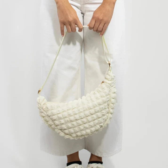 Off-white textured crossbody bag with a crescent shape and light-colored straps.