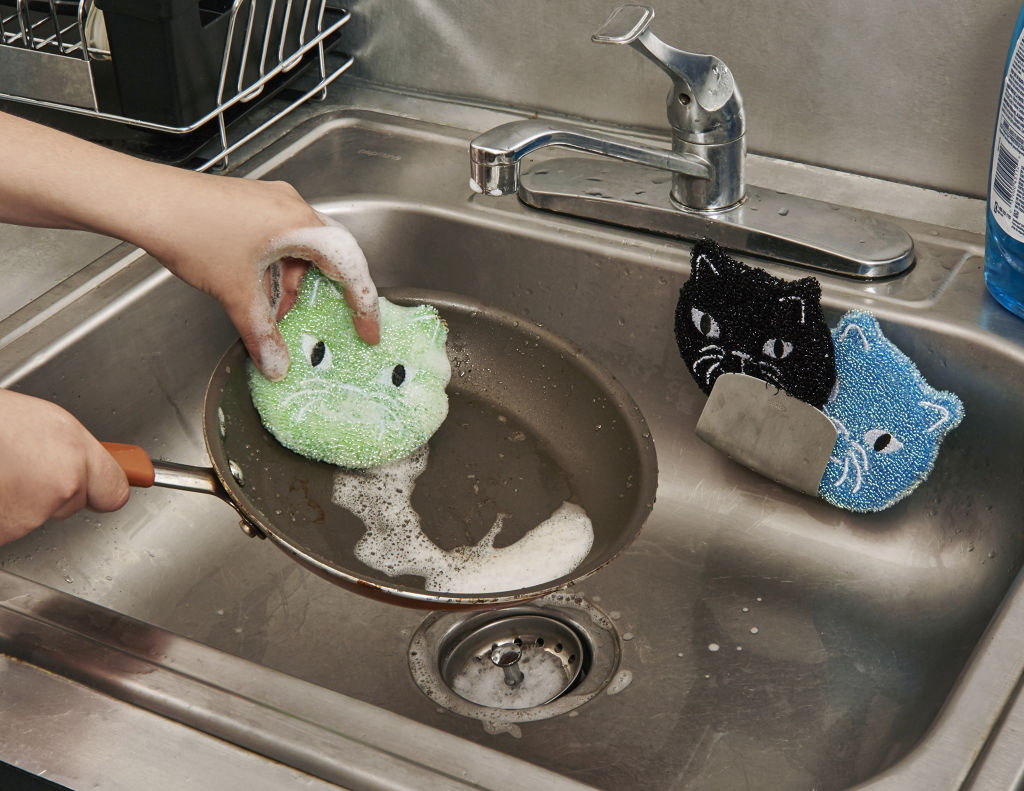 A person washing a green cat-shaped scrubbing sponge in a frying pan.