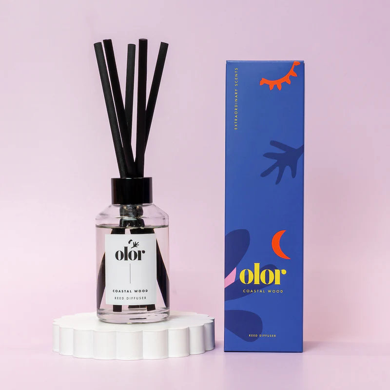 Clear glass reed diffuser bottle with black reeds and black cap;  "olor" label; Coastal Wood scent.  Matching blue box shown.
