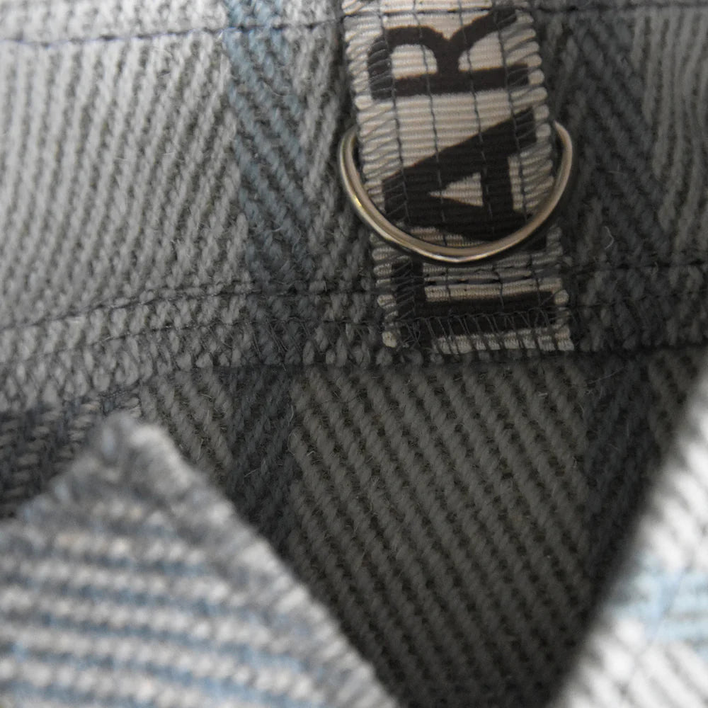 Close-up of a gray and blue plaid fabric with a woven label attached; the label features black lettering and a metal ring.