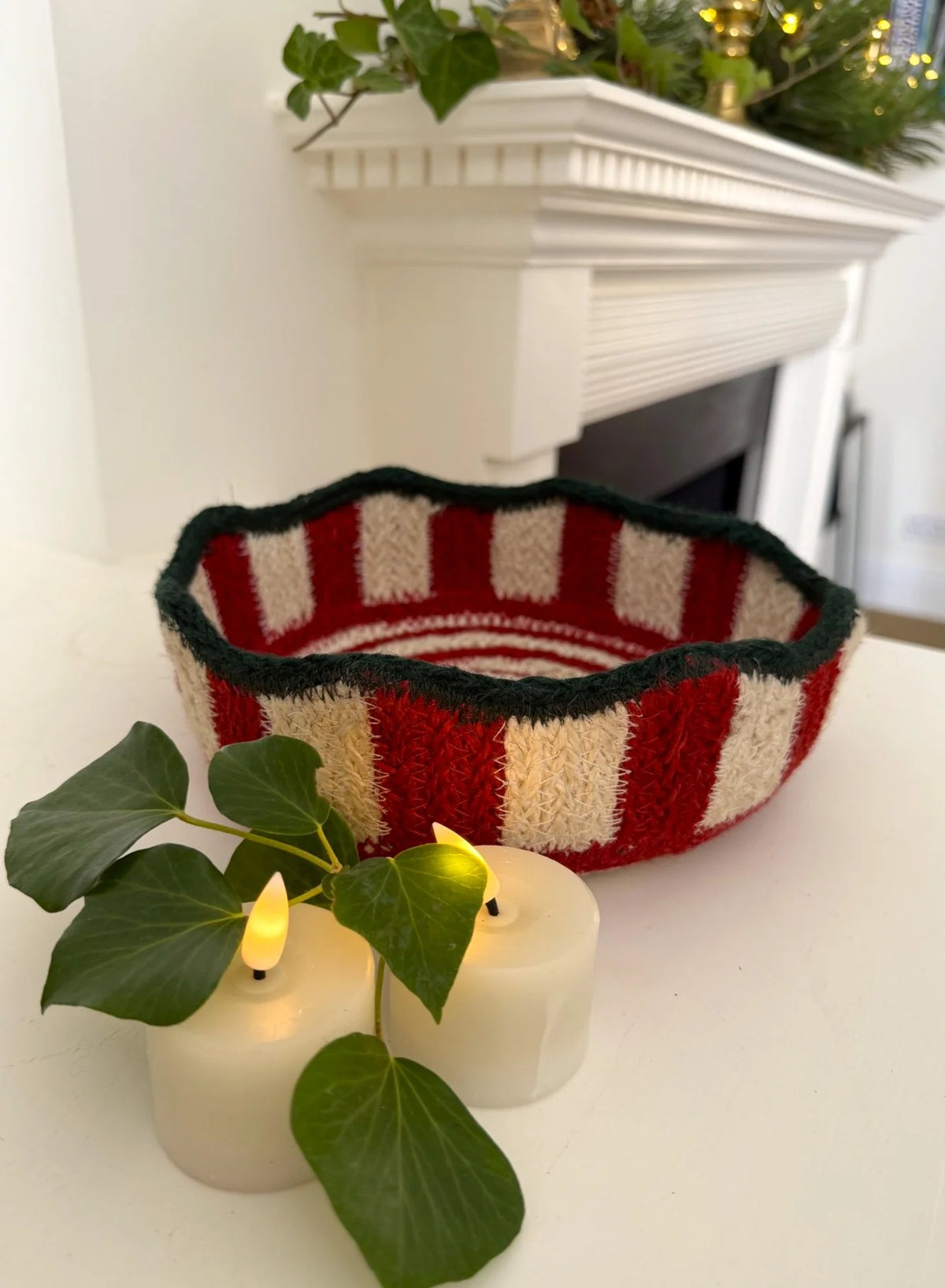 A hand-knit holiday bowl with red and cream stripes and a dark green trim, styled on a white mantelpiece with two lit candles and green ivy in the foreground.