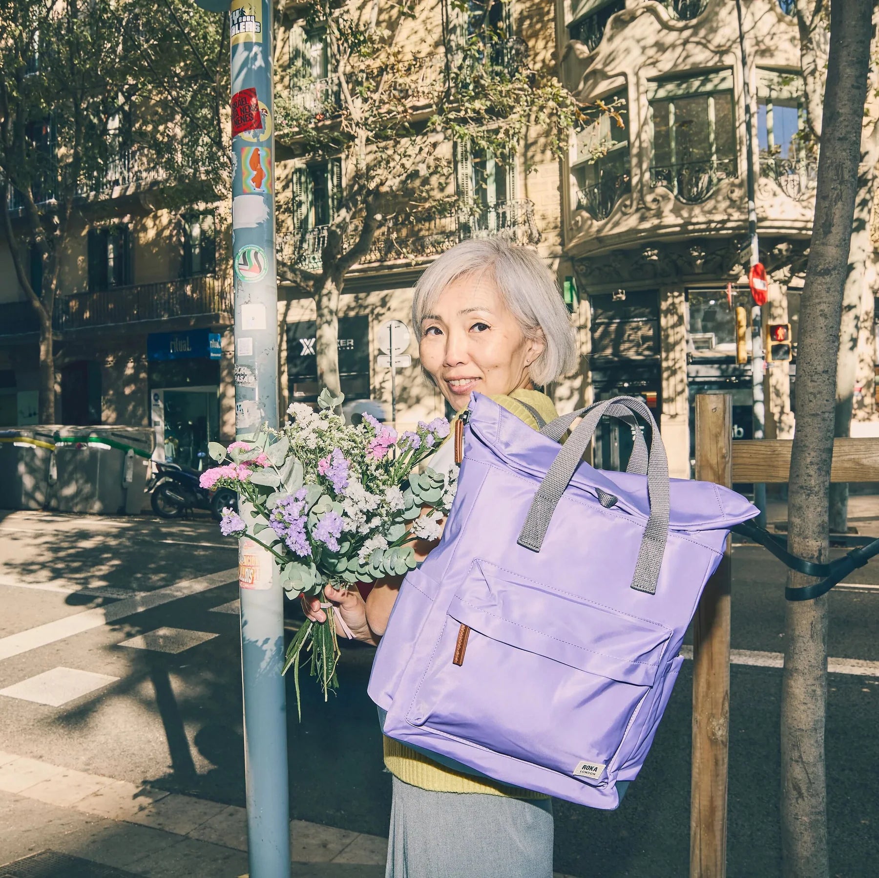 Woman holding a bouquet of flowers and a purple backpack on a city street.