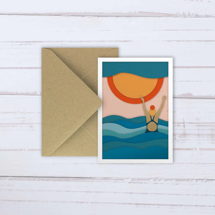 Paper cut-out greeting card depicting a swimmer emerging from the ocean at sunset.