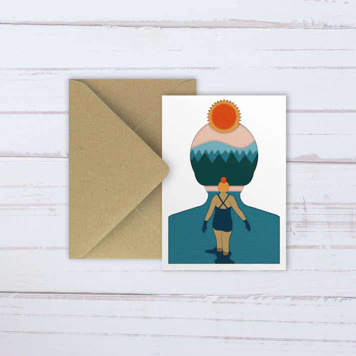 Layered paper art greeting card depicting a woman wading in water, her head replaced by a sun, sky, and mountain landscape.