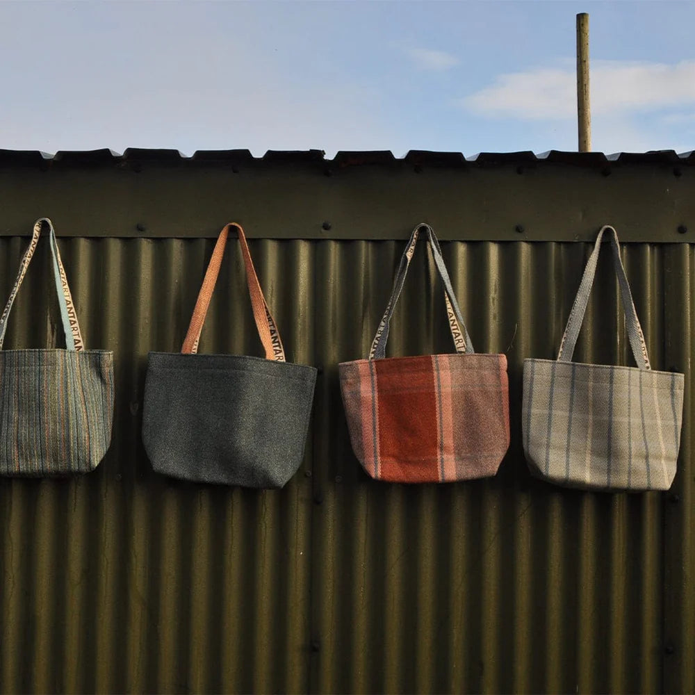 Four small tote bags in various woven fabrics hang against a corrugated metal wall.