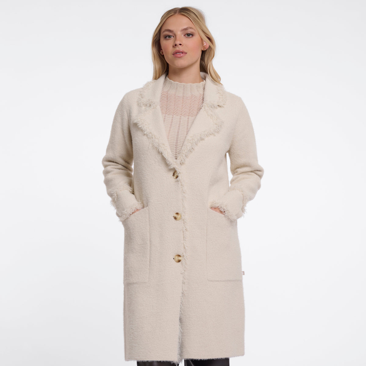A woman modeling a long cream-colored textured coat with fuzzy trim and gold buttons, paired with dark brown wide-leg trousers, against a plain white studio background.